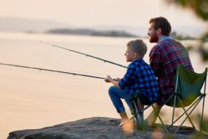Side view portrait of father and son sitting together on rocks fishing with rods in calm lake waters with landscape of setting sun, both wearing checkered shirts, shot from behind tree